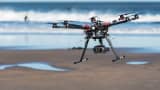 An octocopter photographic drone, equipped with a video camera, hovers over a beach.