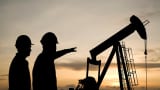 Two workers stand before the backdrop of an oil pump, while silhouetted against the sunset.