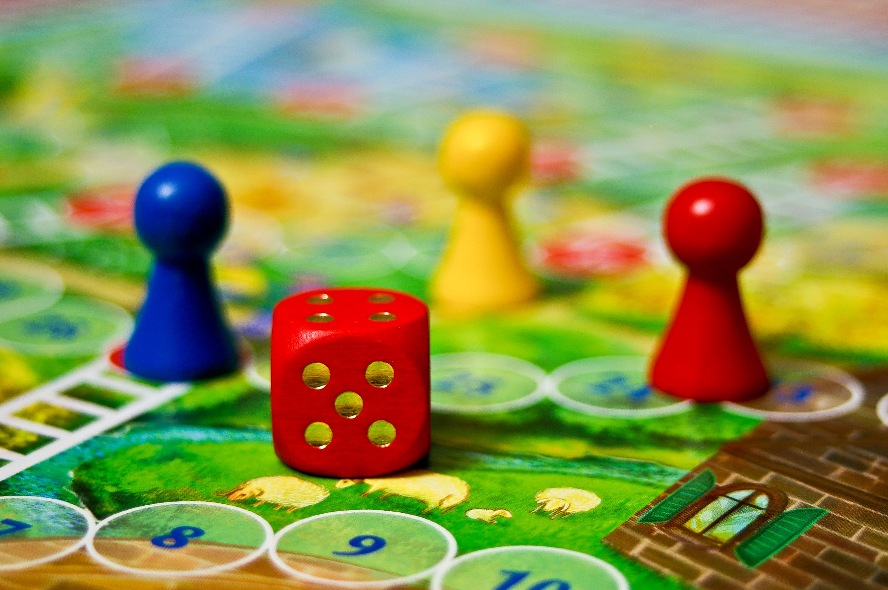  A red game die sits in the foreground of a colorful board game with blue, yellow, and red player pieces.