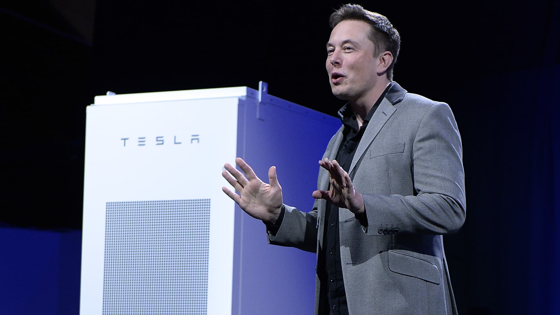 Elon Musk, CEO of Tesla, with a Powerpack unit the background unveils suit of batteries for homes, businesses, and utilities.