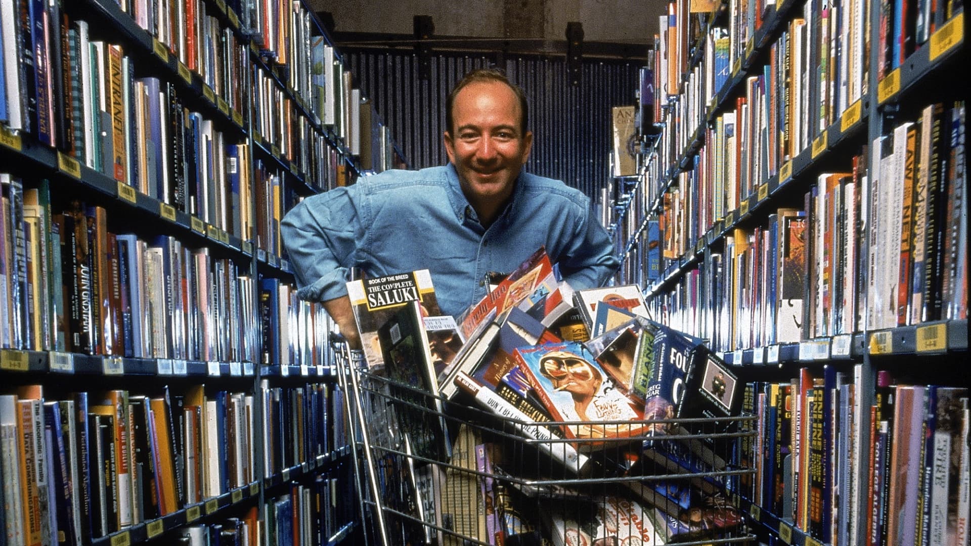 Portrait of American businessman and Amazon.com CEO Jeff Bezos poses in an aisle of bookshelves with a shopping cart full of books and compact discs, Seattle, Washington, September 1998.