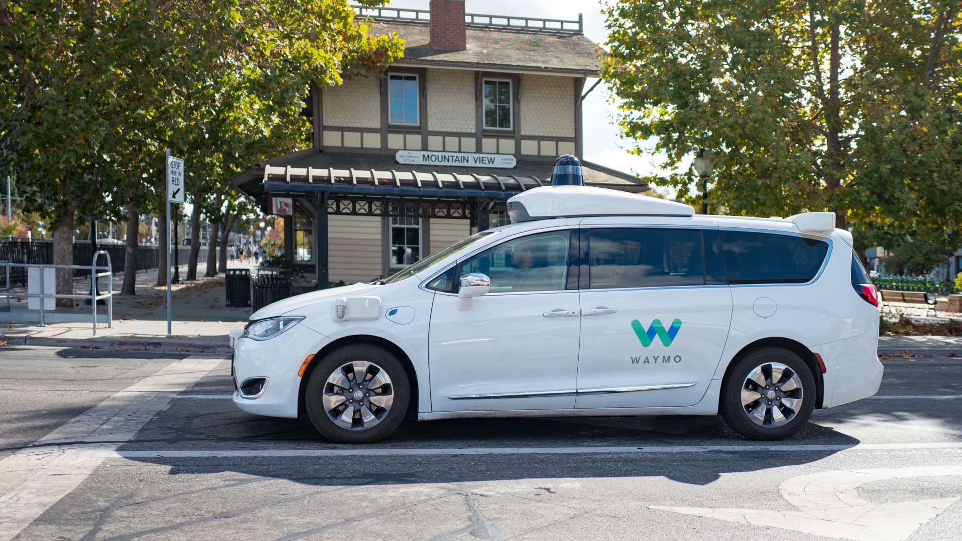 Close-up of self driving minivan, with LIDAR and other sensor units and logo visible, part of Google parent company Alphabet Inc, driving past historic railroad station with sign reading Mountain View, in the Silicon Valley town of Mountain View, California, with safety driver visible, October 28, 2018.