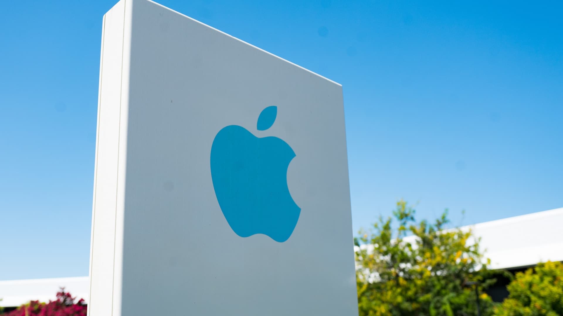 Close-up of blue logo on sign with facade of headquarters buildings in background near the headquarters of Apple Computers in the Silicon Valley, Cupertino, California, August 26, 2018.