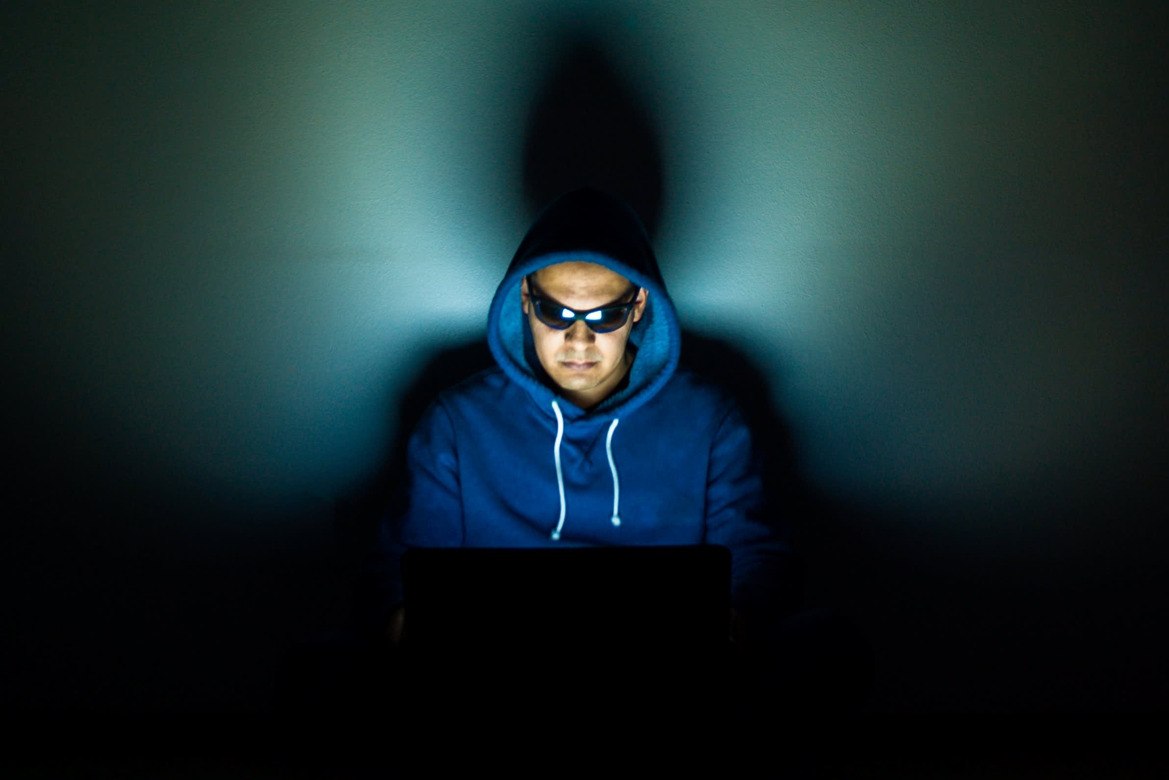 trope photo of a ‘hacker’ in hoodie and sunglasses lit by his laptop backlight