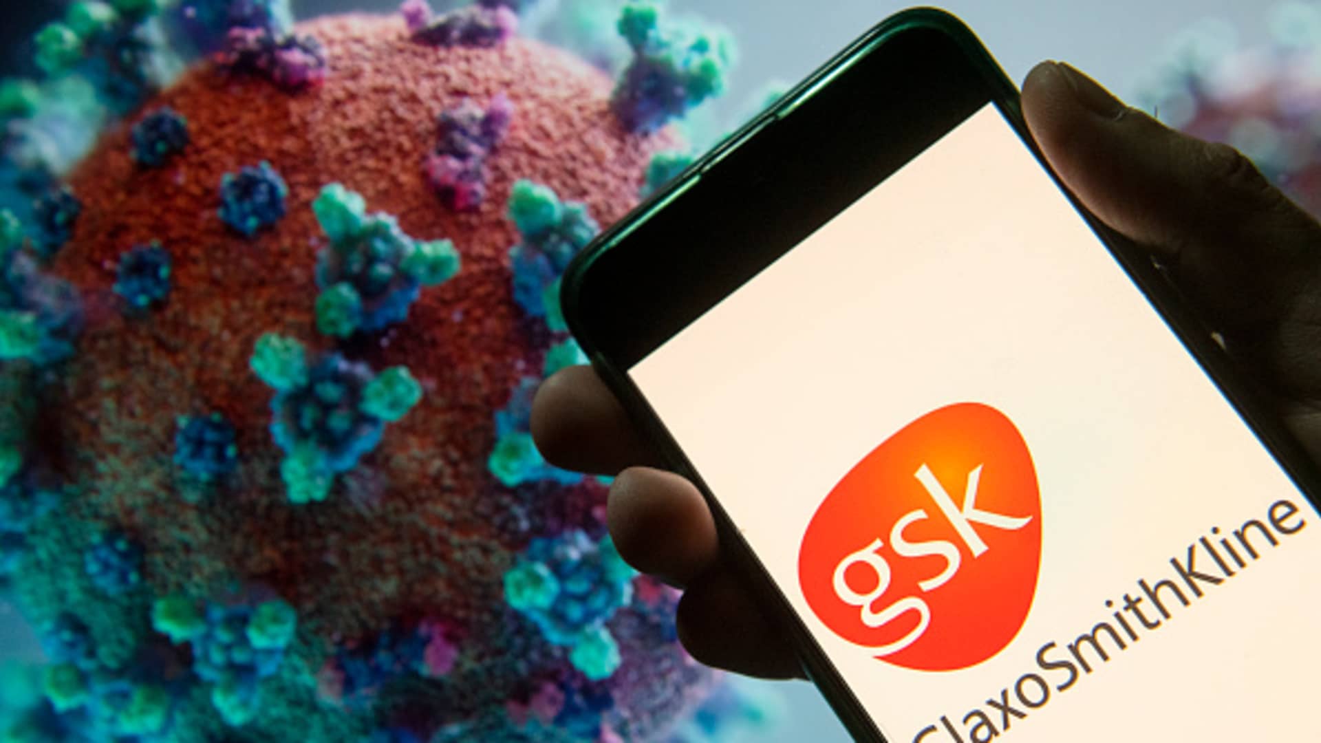 In this photo illustration the British multinational pharmaceutical company GlaxoSmithKline (GSK) logo seen displayed on a smartphone with a computer model of the COVID-19 coronavirus on the background.