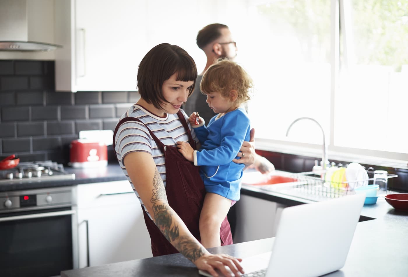 Dad washing up while mum looks after daughter and uses laptop