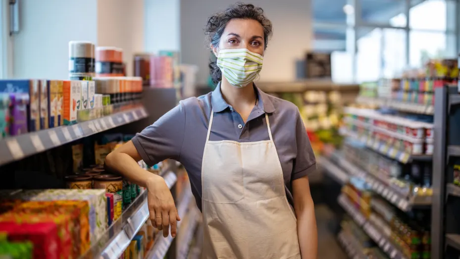 Portrait of a woman working in supermarket wearing a face mask looking at camera. Store assistance with face mask and apron leaning to a rack in grocery store.