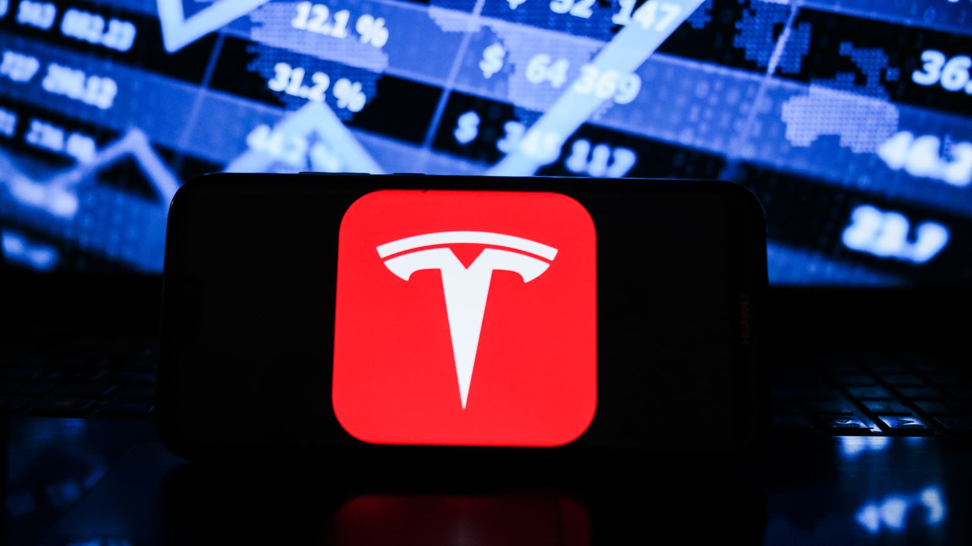 In this photo illustration a Tesla logo seen displayed on a smartphone with the stock market graphic in the background.