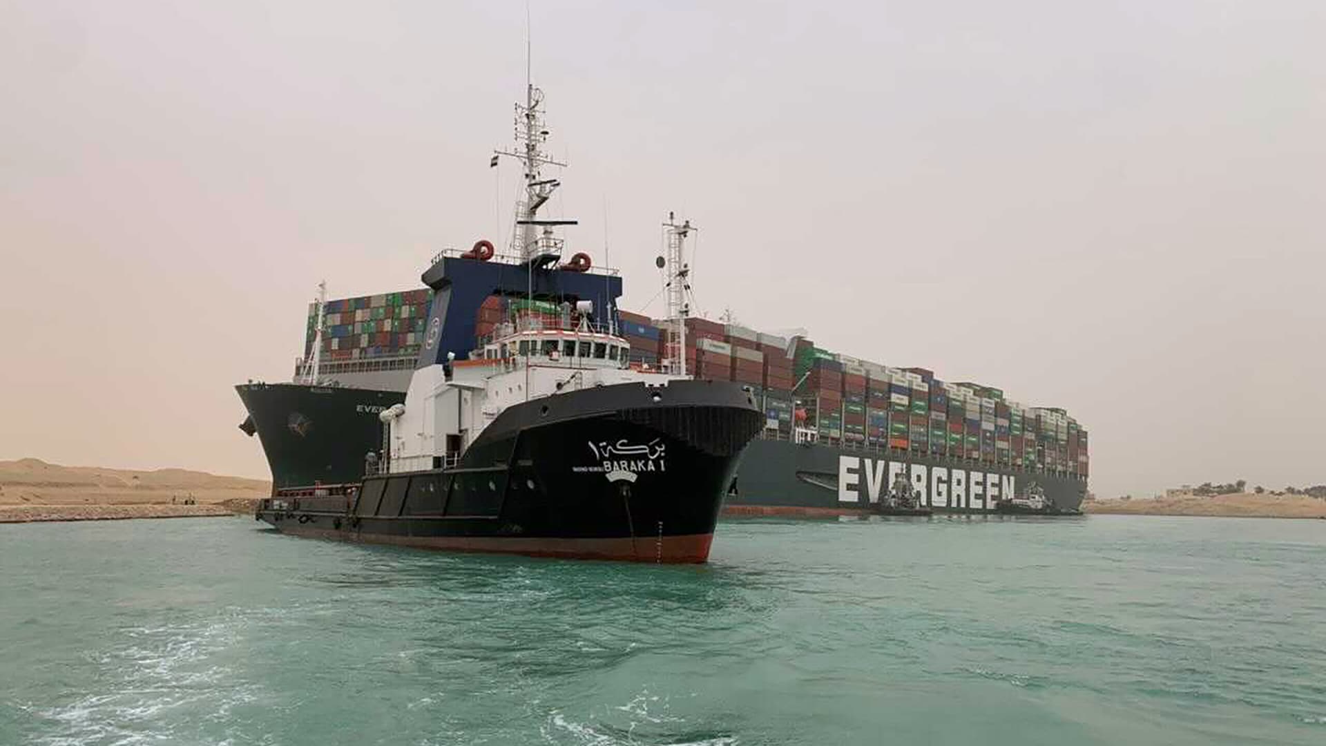 In this photo released by the Suez Canal Authority, a boat navigates in front of a massive cargo ship, named the Ever Given, rear, sits grounded Wednesday, March 24, 2021, after it turned sideways in Egypt's Suez Canal, blocking traffic in a crucial East-West waterway for global shipping.