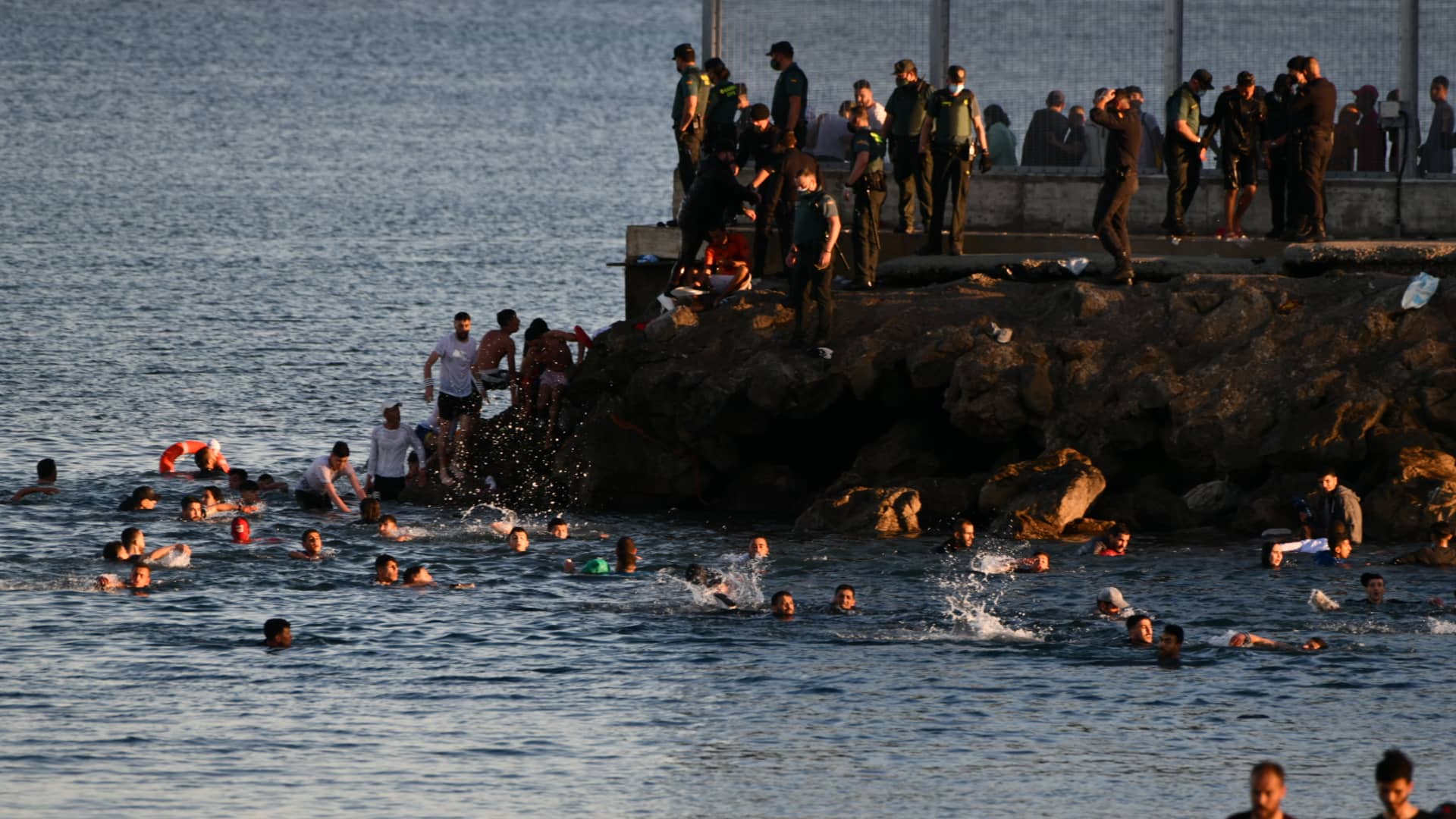 Migrants swim towards the beach of Tarajal from Morocco, as others already on the Spanish side walk along the beach on May 17, 2021 in Ceuta, Spain.
