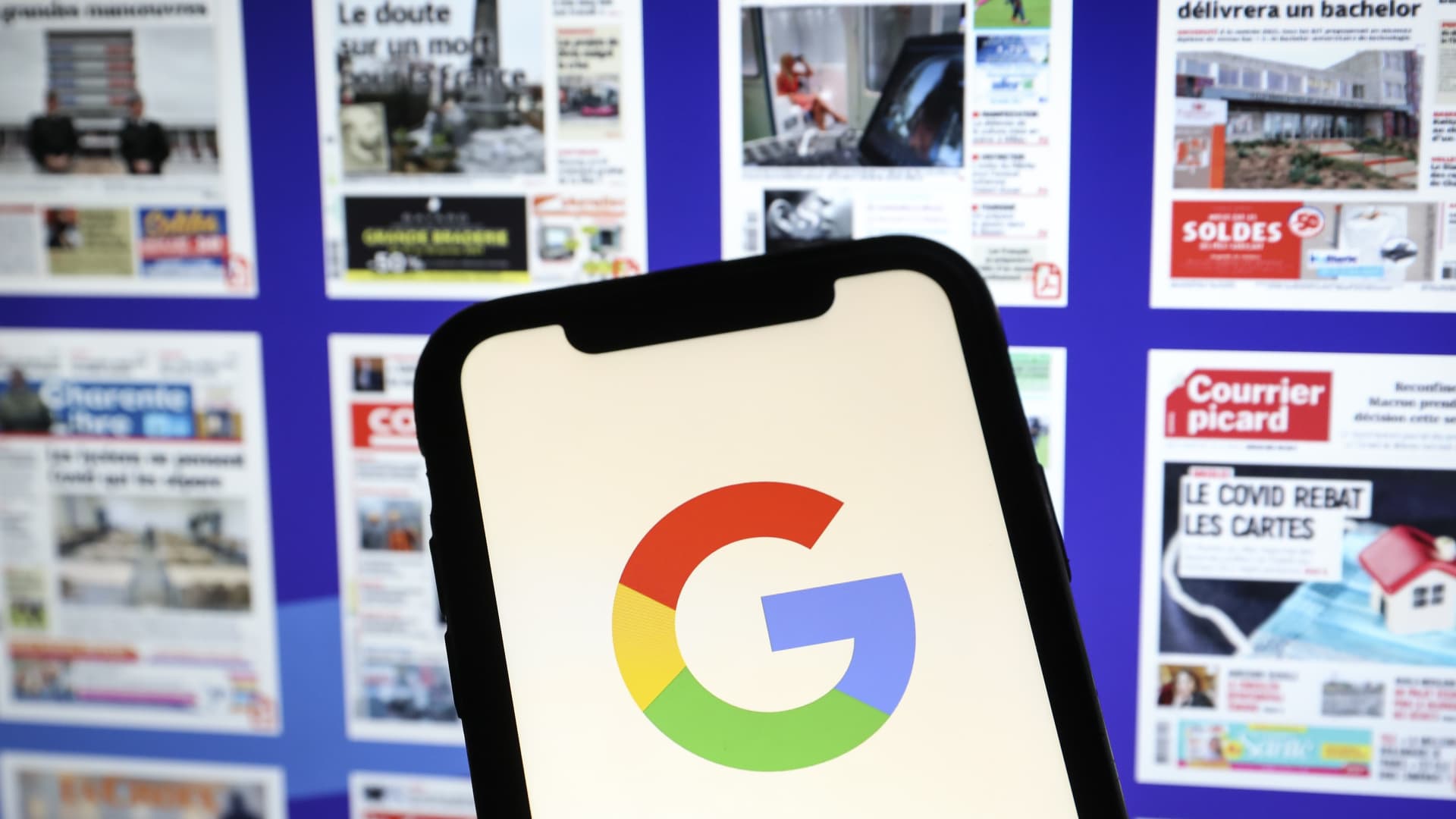 The Google logo displayed on a smartphone with the front pages of several newspapers pictured in the background.