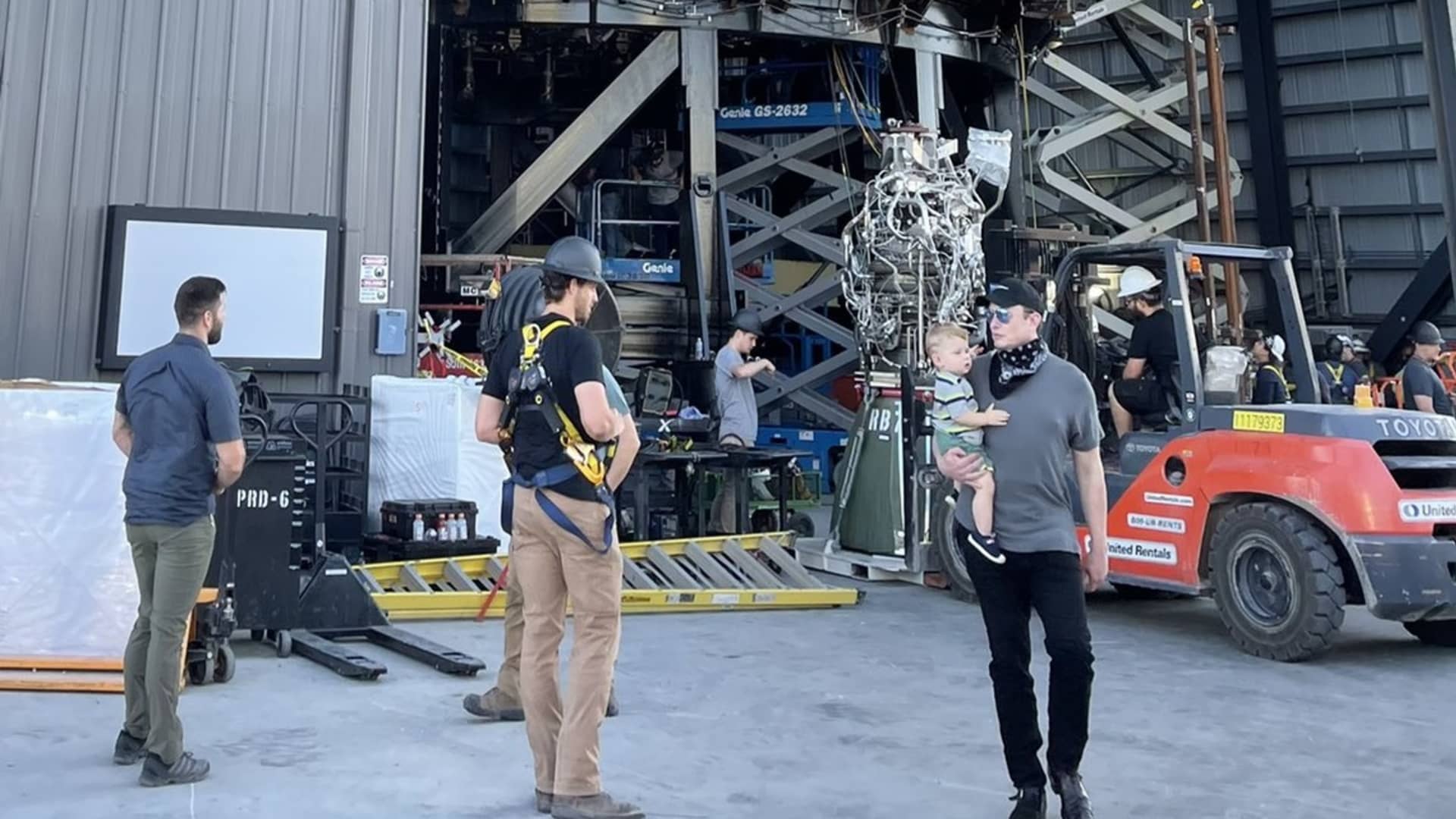 Musk, carrying his son, walks near a Raptor rocket engine being installed in SpaceX's Super Heavy Booster 4.