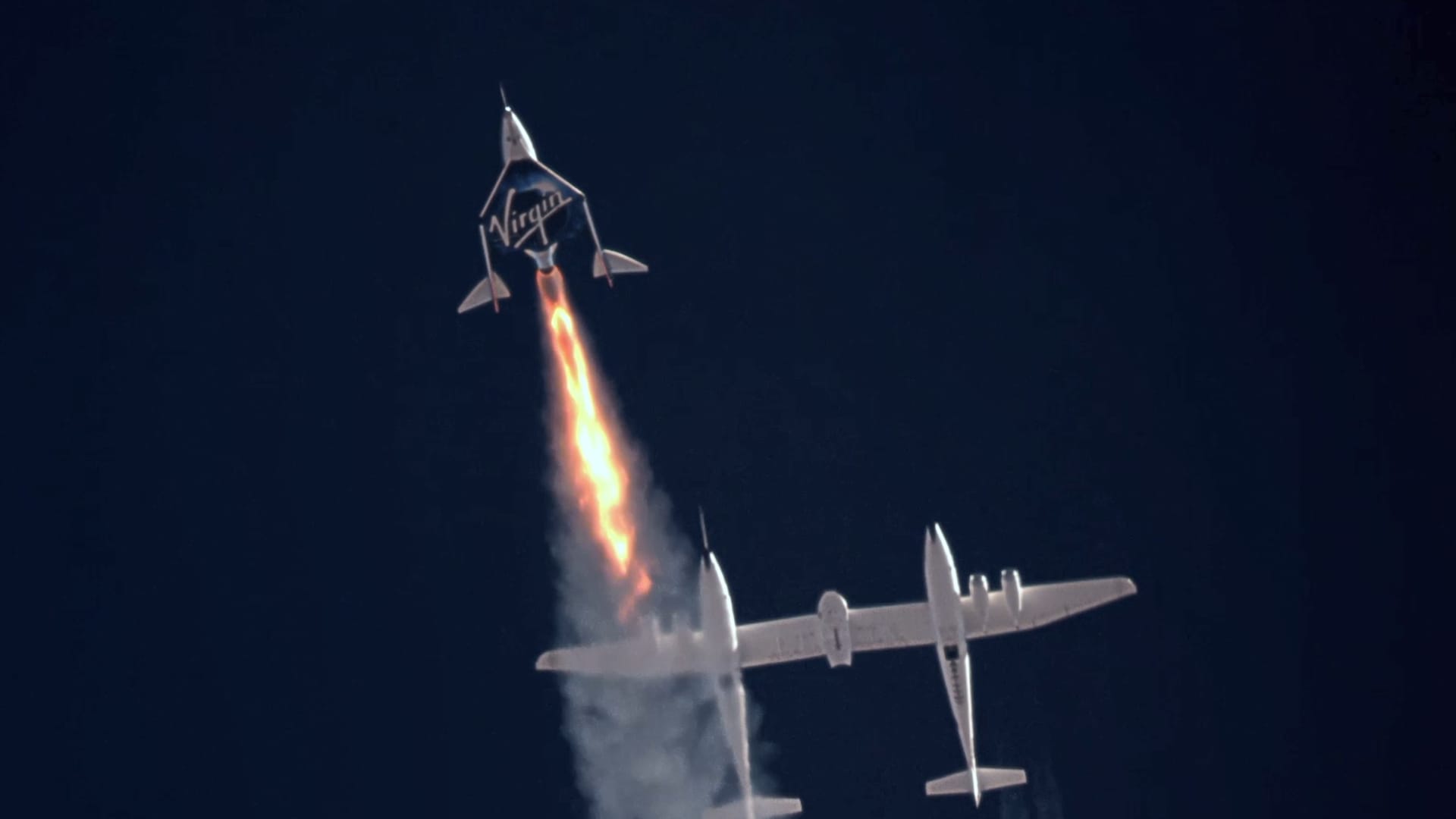 Carrier aircraft VMS Eve is seen in the background shortly after releasing VSS Unity, which is firing its engine and acclerating during the company's fourth spaceflight test, Unity 22, carrying founder Richard Branson on July 11, 2021.