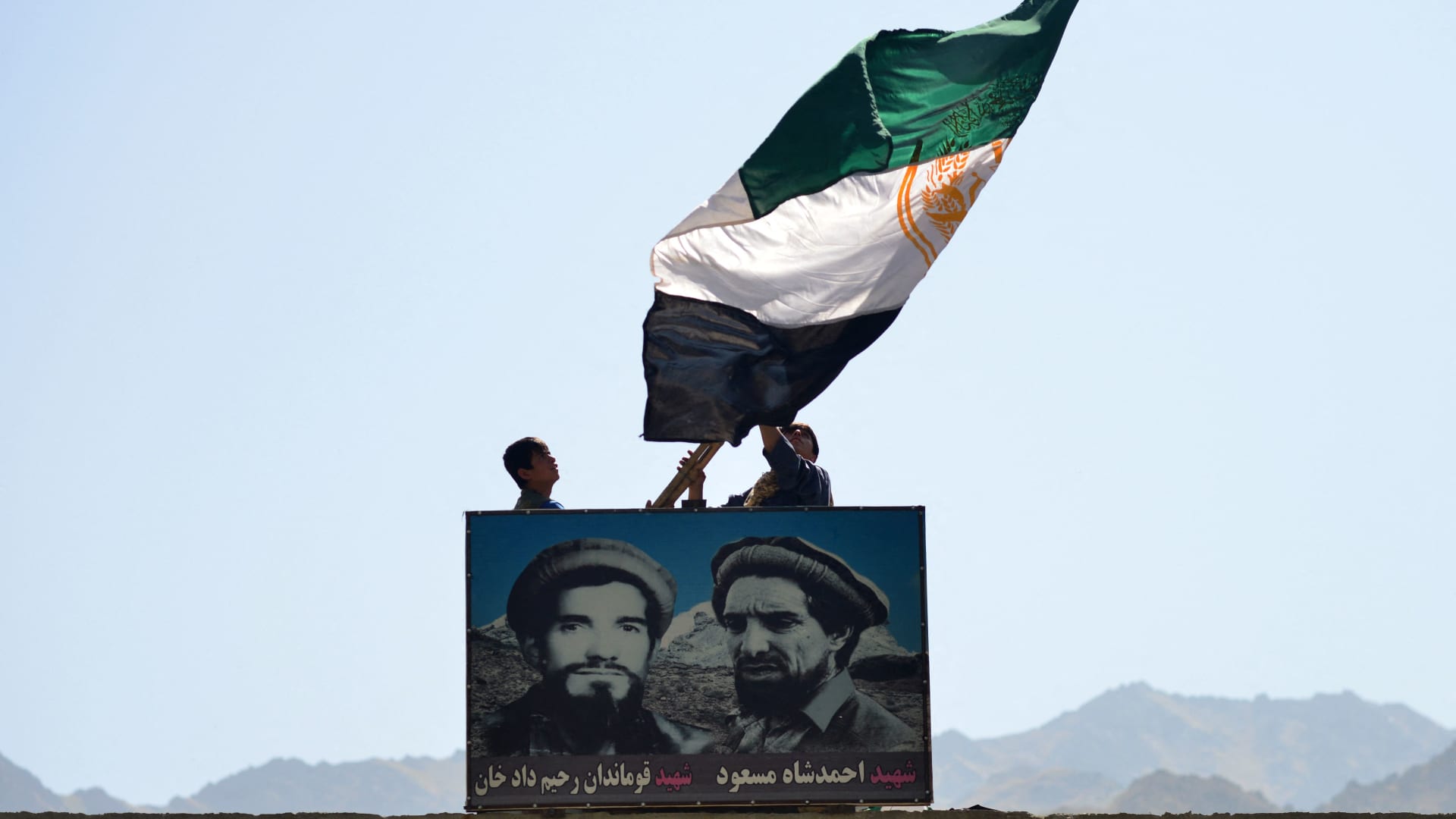 Afghan men wave a flag above the portrait of late Afghan commander Ahmad Shah Massoud (R) in Paryan district of Panjshir province on August 23, 2021, as the Taliban said their fighters had surrounded resistance forces holed up in the valley, but were looking to negotiate rather than take the fight to them.