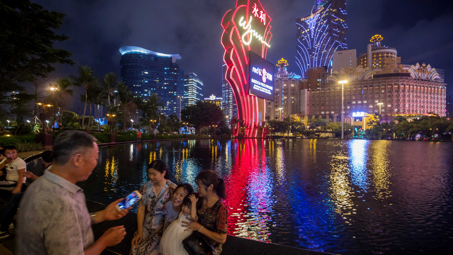 The Wynn Resorts logo stands illuminated as people sit by the fountain at the Wynn Macau casino resort in Macau, China, on Tuesday, July 24, 2018.