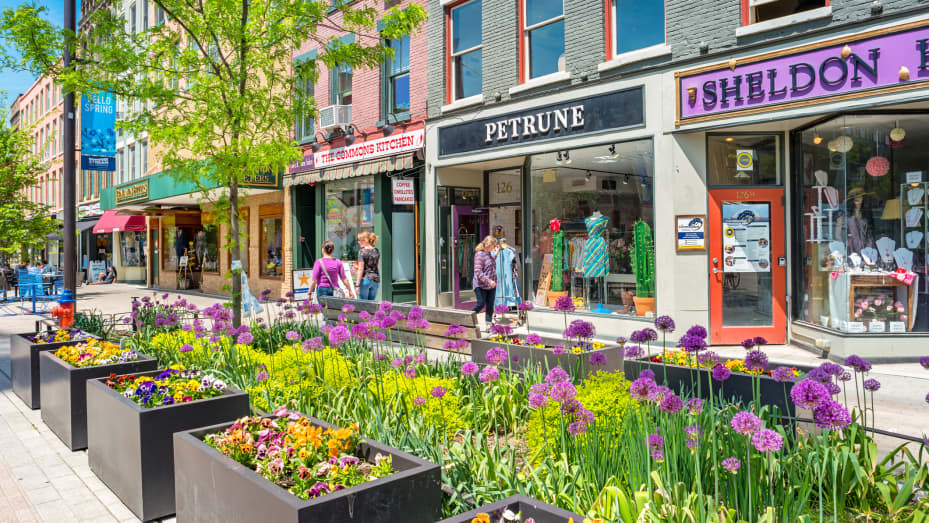 People walk past colorful stores in a pedestrian area of downtown Ithaca, New York on a sunny day.