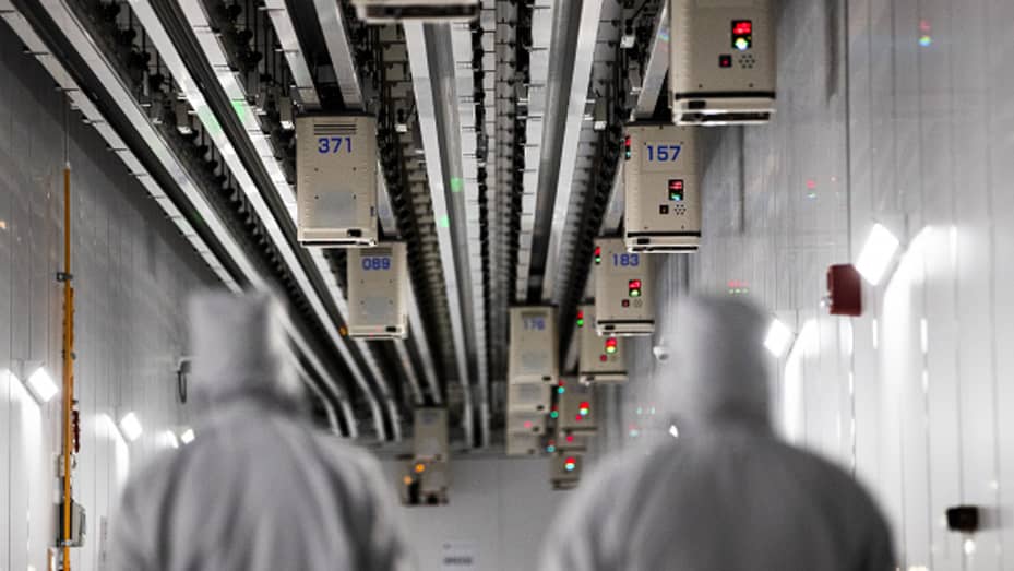 Employees wearing cleanroom suits walk beneath Automated Material Handling Systems (AMHS) vehicle robots moving along tracks on the ceiling inside the GlobalFoundries semiconductor manufacturing facility in Malta, New York, U.S., on Tuesday, March 16, 2021.