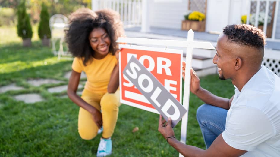 A young heterosexual Black couple puts a "sold" sticker on the "For sale" sign in front of a house.