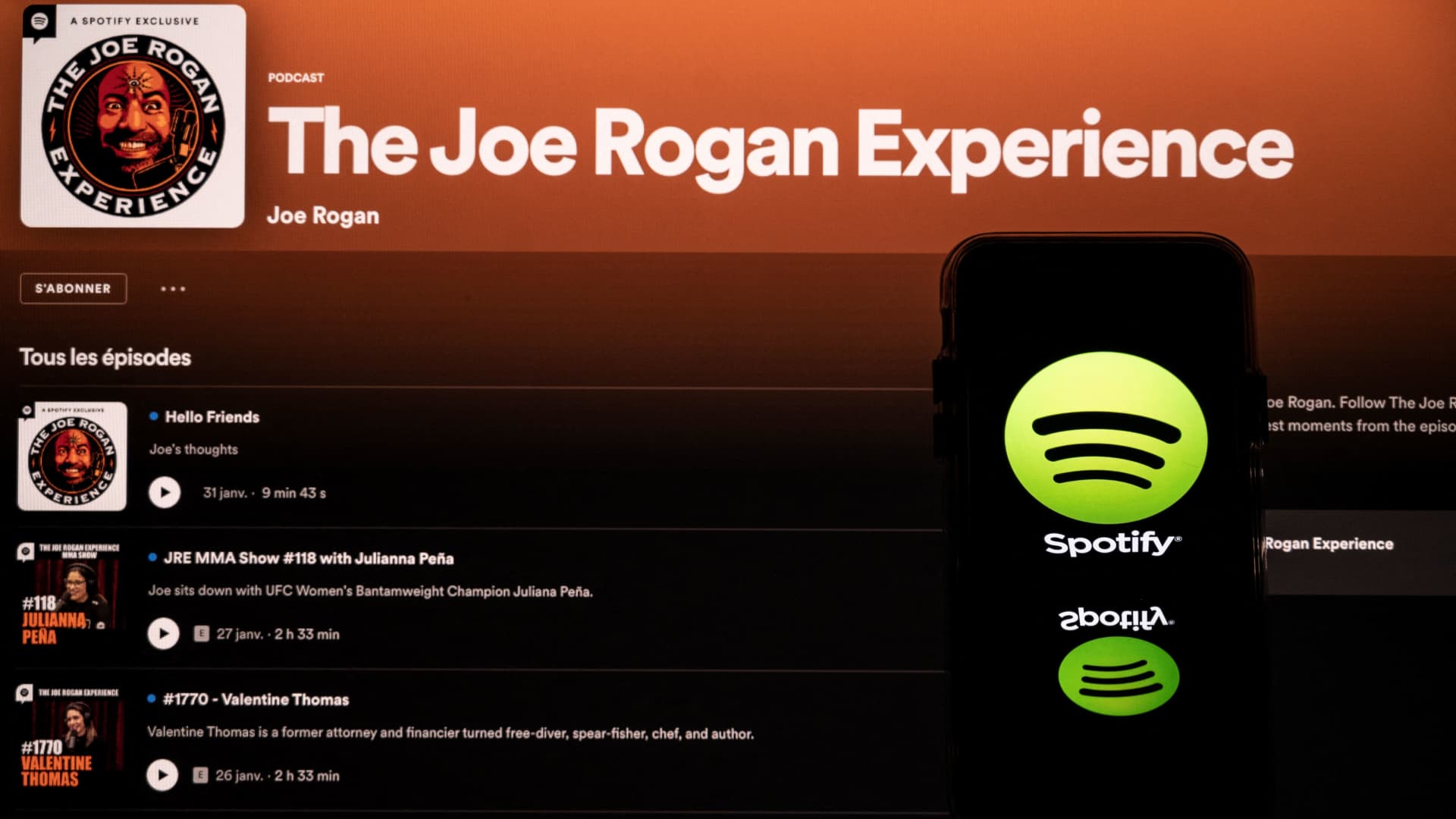 This picture taken in Toulouse, southwestern France, on January 31, 2022 shows a screen displaying the Joe Rogan podcast and a smartphone displaying the Spotify's logo.