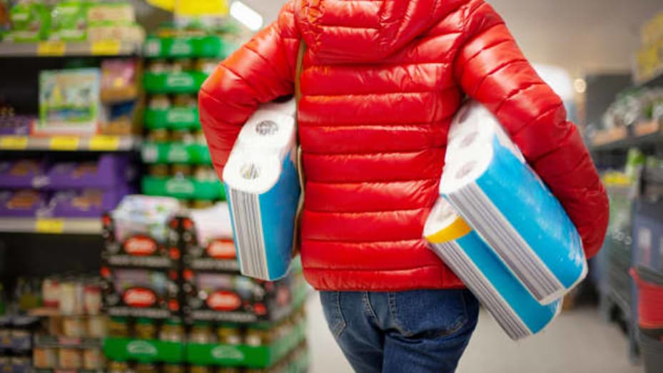 Shopper in grocery store wearing red puffy coat and holding three packs of toilet paper.