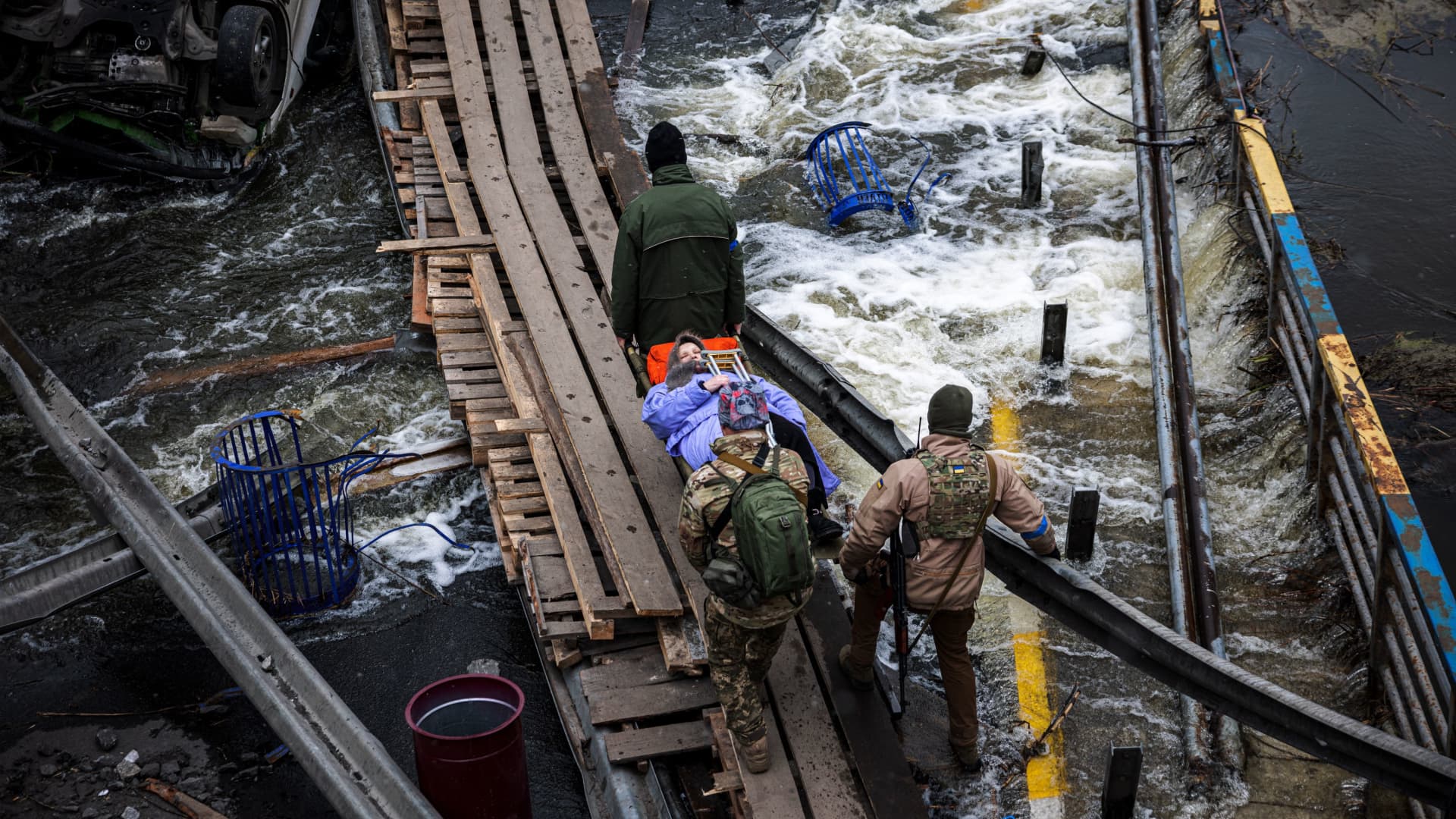 Ukrainian servicemen carry an elderly woman on a stretcher on a makshift pathway to cross a river next to a destroyed bridge as people flee the city of Irpin, northwest of Kyiv, on March 13, 2022.