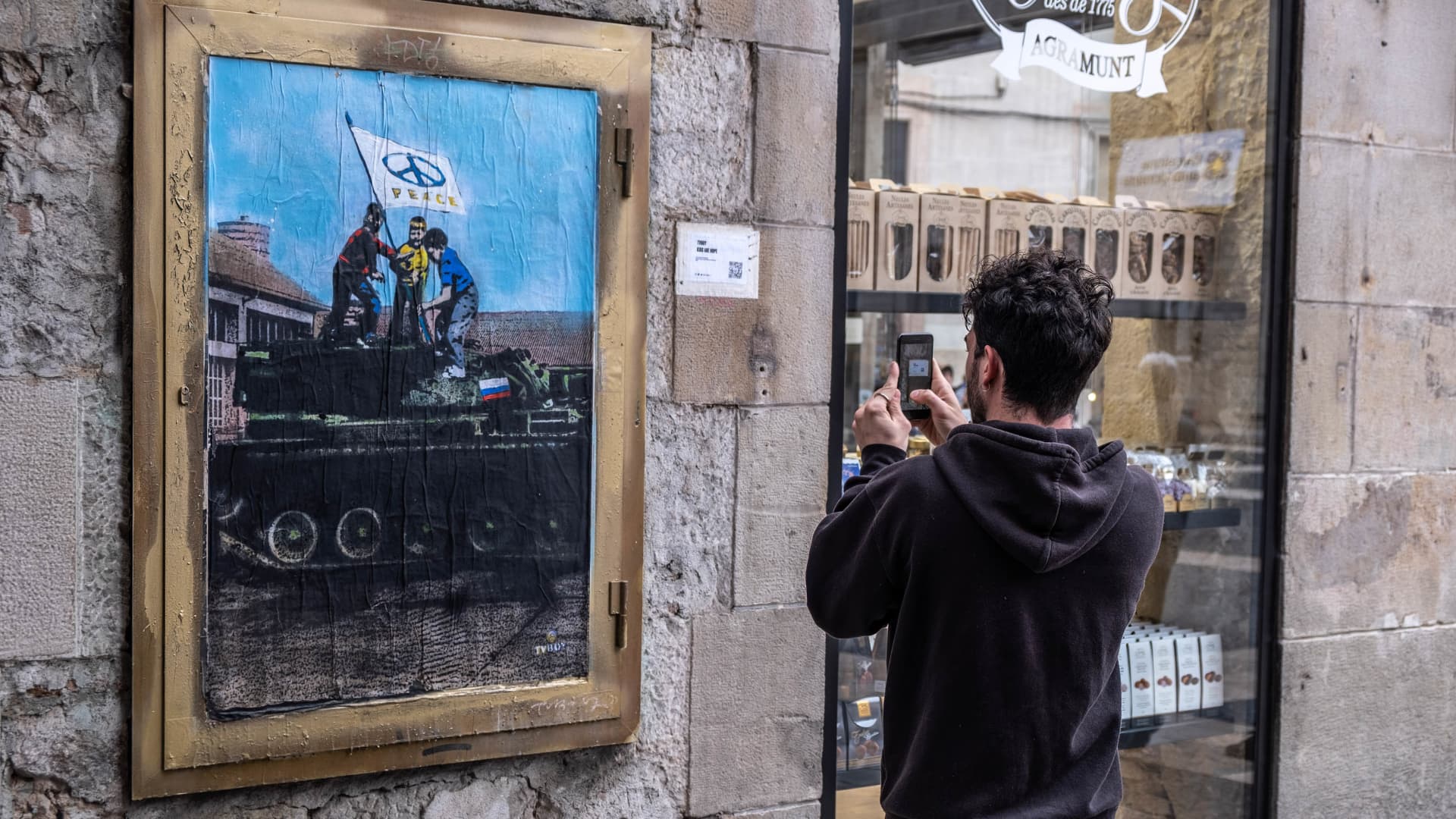 A passer-by is seen taking photos of artist TvBoy's new collage for peace in Ukraine. TvBoy, the Italian artist living in Barcelona, installs a new collage on the war in Ukraine in Plaza de Sant Jaume, representing three children installing a flag of peace on a Russian tank.
