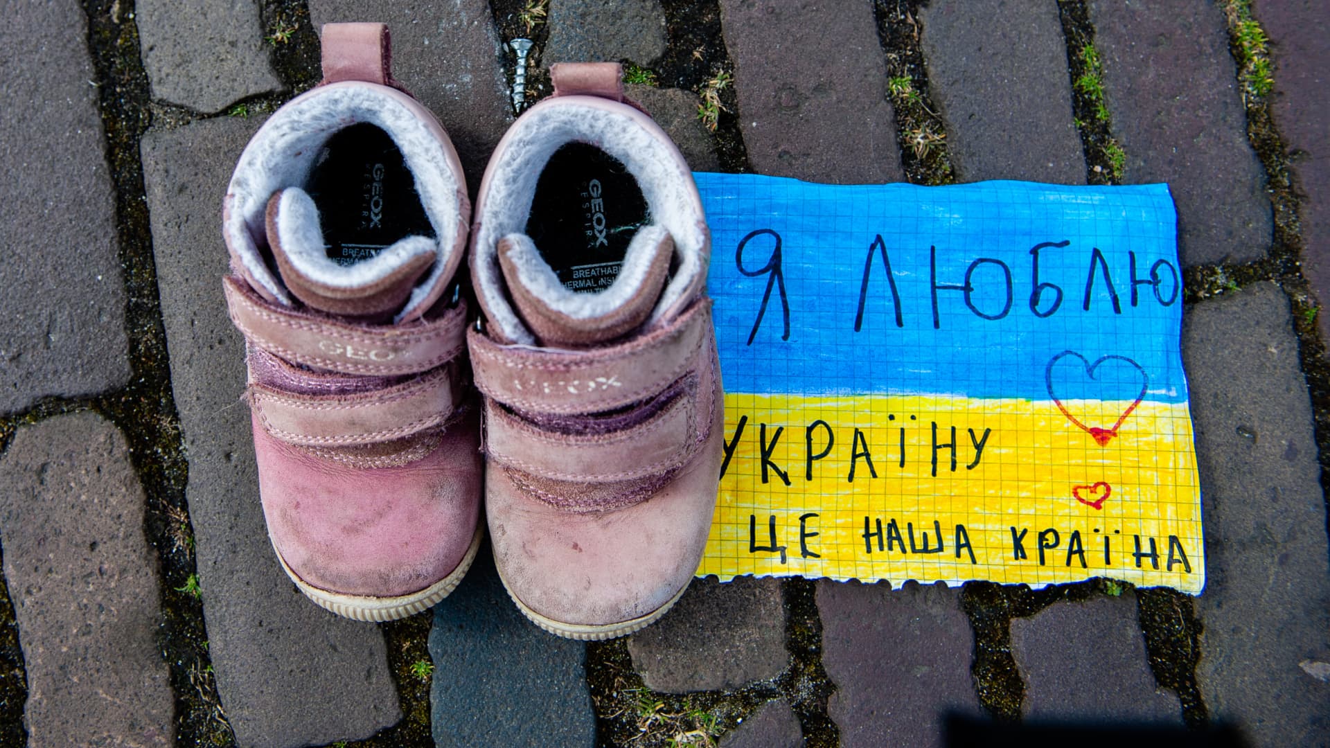 A couple of children's shoes is on the floor close to a placard in support of Ukraine, A woman is putting children's shoes on the floor, as a part of a Ukrainian art installation to draw attention to the killings of civilians and in particular children during the war in Ukraine. The Hague, on April 2nd, 2022.