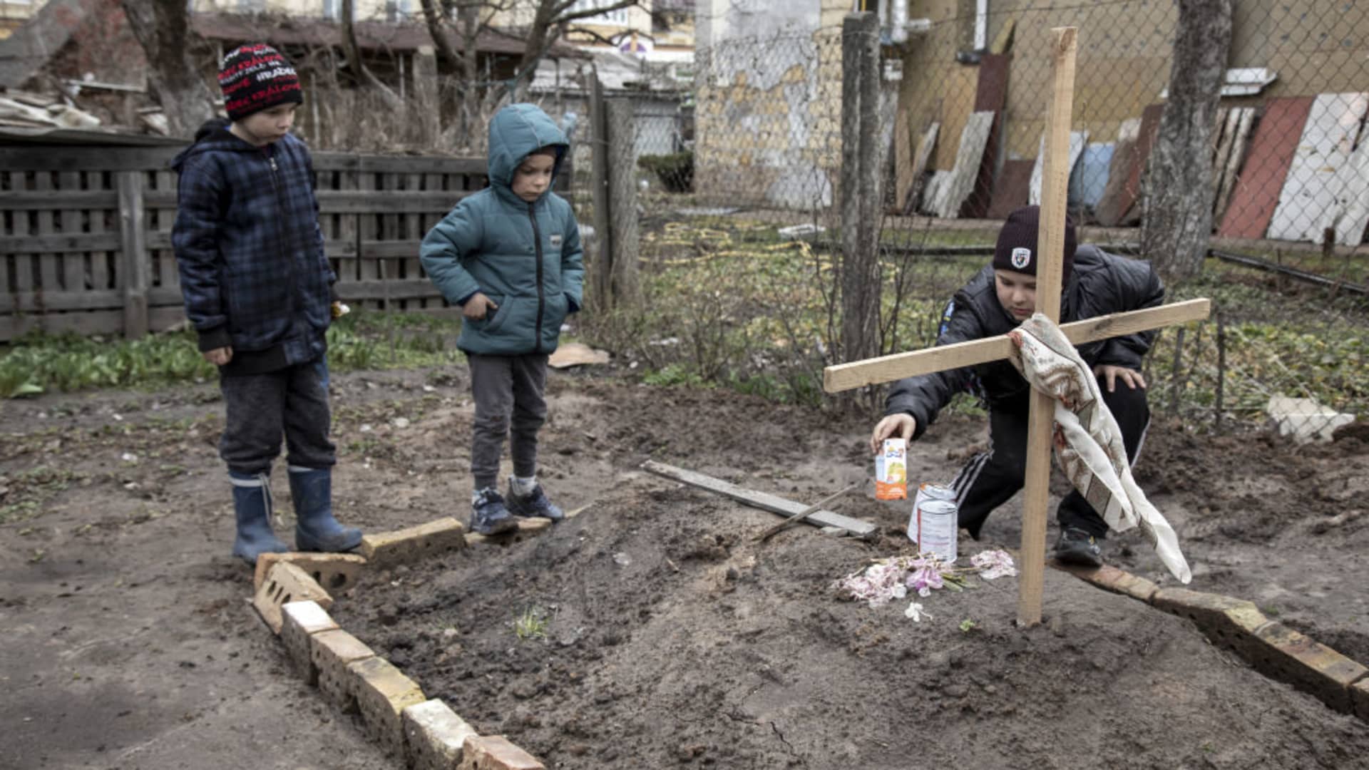 A young boy gives an offering of food to his mother's grave as his younger brother and a neighbor stand next to it, in the town of Bucha, on the outskirts of Kyiv, after the Ukrainian army secured the area following the withdrawal of the Russian army from the Kyiv region on previous days, Bucha, Ukraine, April 4th, 2022.