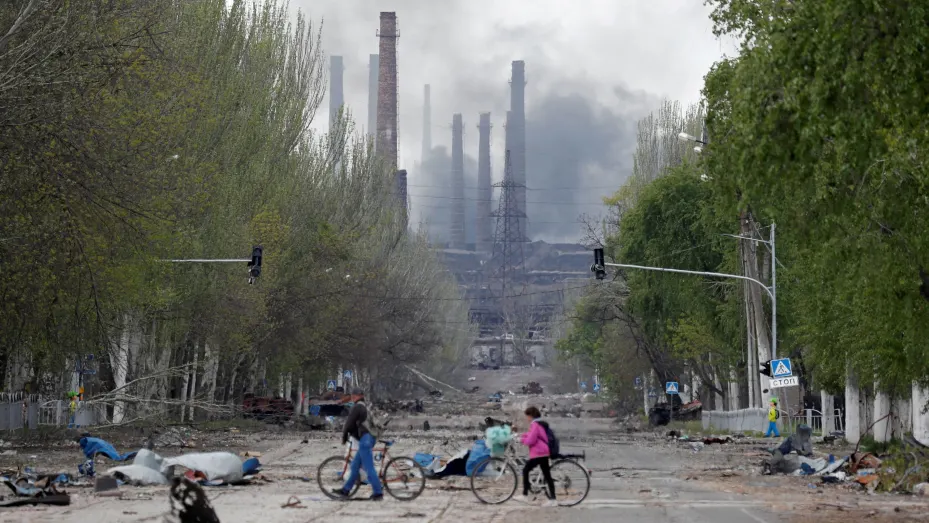 People walk their bikes across the street as smoke rises above a plant of Azovstal Iron and Steel Works during Ukraine-Russia conflict in the southern port city of Mariupol, Ukraine May 2, 2022. REUTERS/Alexander Ermochenko     TPX IMAGES OF THE DAY