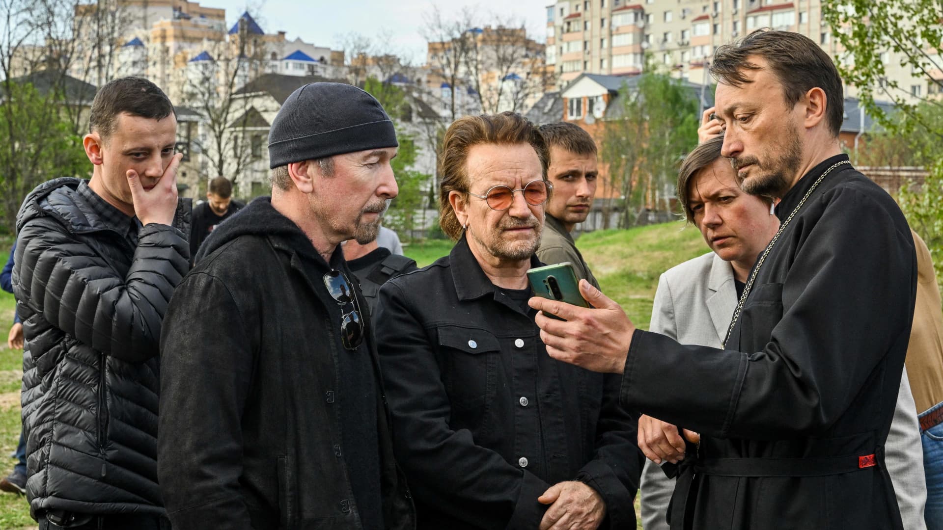Andrii Holovine, priest of the Church of St. Andrew Pervozvannoho All Saints (R) shows Bono (C) (Paul David Hewson), activist and front man of the Irish rock band U2 and guitarist David Howell Evans aka 'The Edge' (L) images on his cell phone visiting the site of a mass grave by the church in the Ukrainian town of Bucha, near Kyiv on May 8, 2022.