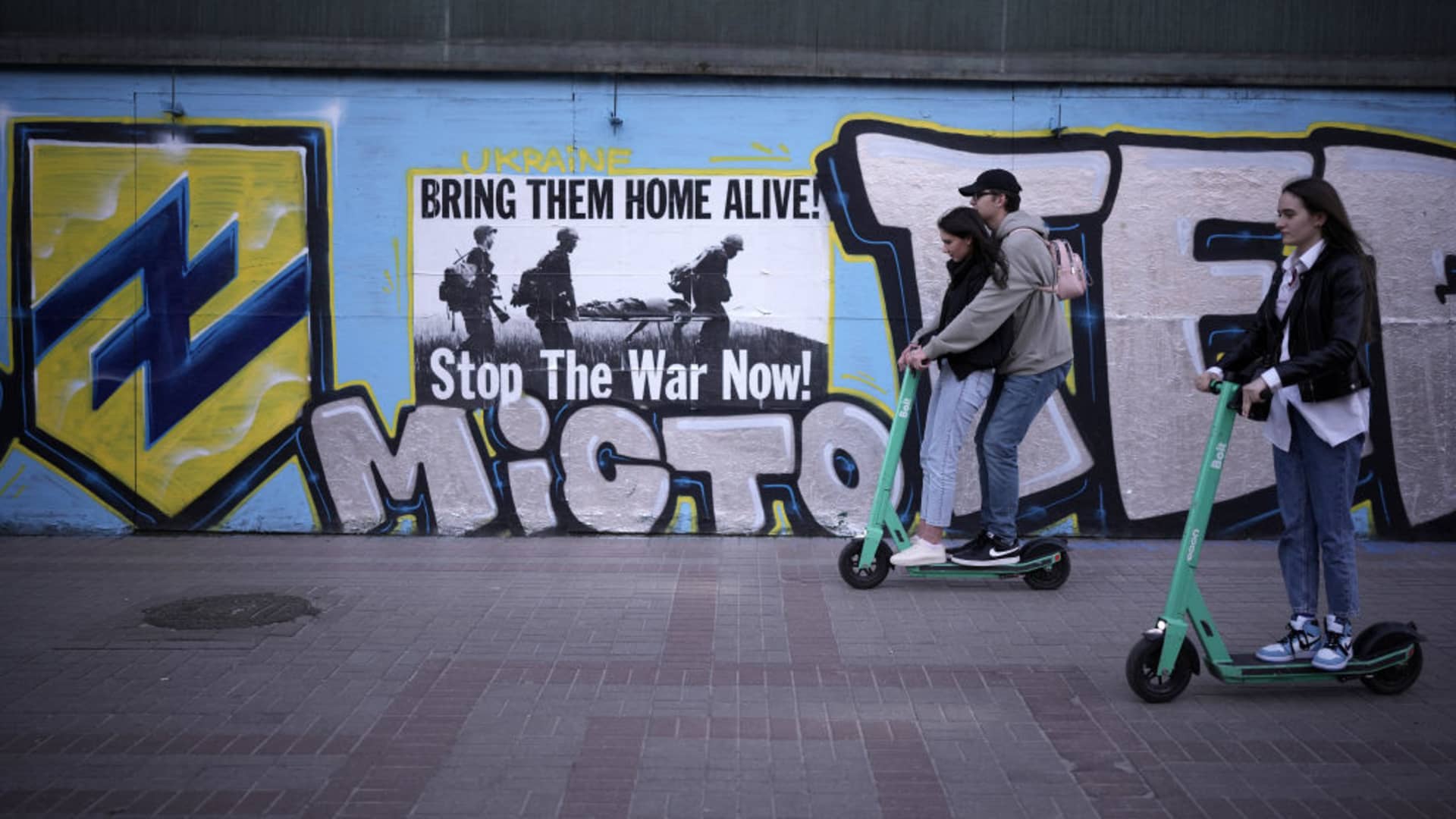 Young people ride their e-scooters past a 'stop the war' poster in central Kyiv on May 19, 2022 in Kyiv, Ukraine.