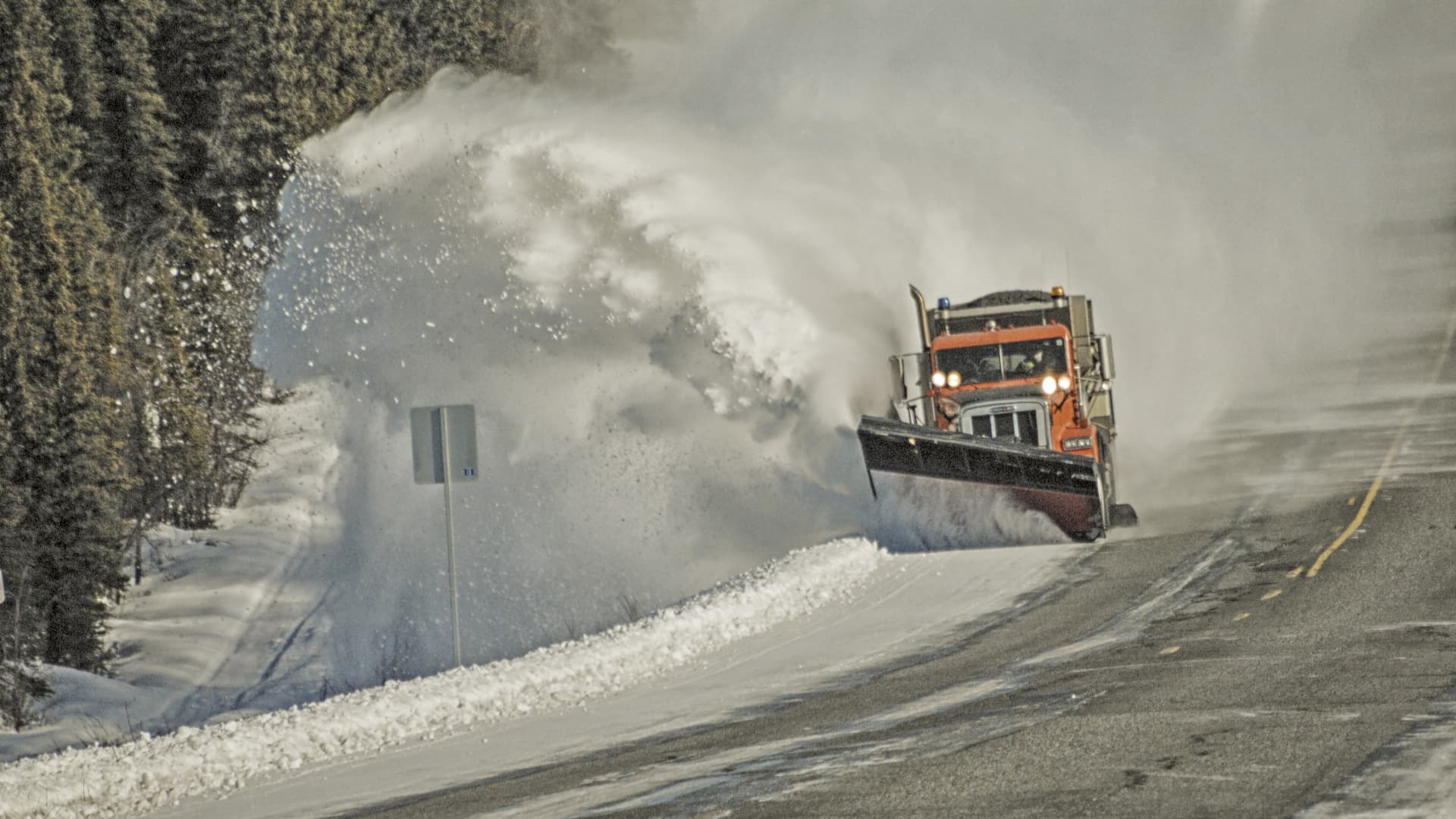 Orange snowplow makes its way down the highway, clearing the snow as it goes.Picture was taken in Interior Alaska on a bright day in winter.