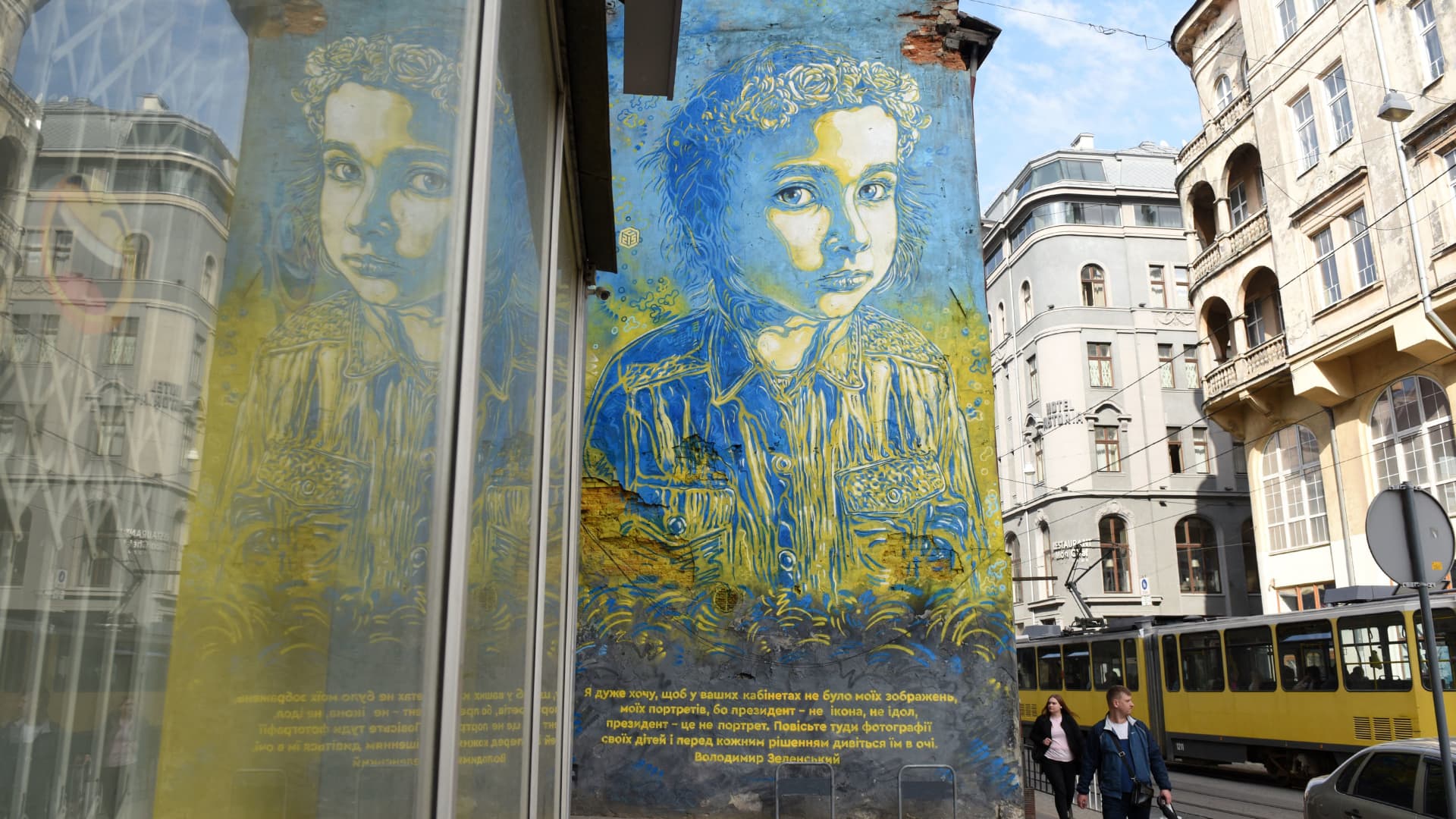In this photograph taken on May 13, 2022, pedestrians walk past a fresco by street-artist Christian Guemy, known as C215, in the western Ukrainian city of Lviv. - The frescos depicting a Ukrainian young girl with a quote attributed to Ukrainian President Volodymir Zelensky I really don't want my photos in your offices, because I am neither a god nor an icon, but rather a servant of the Nation. Instead, hang pictures of your children and look at them whenever you want to make a decision.