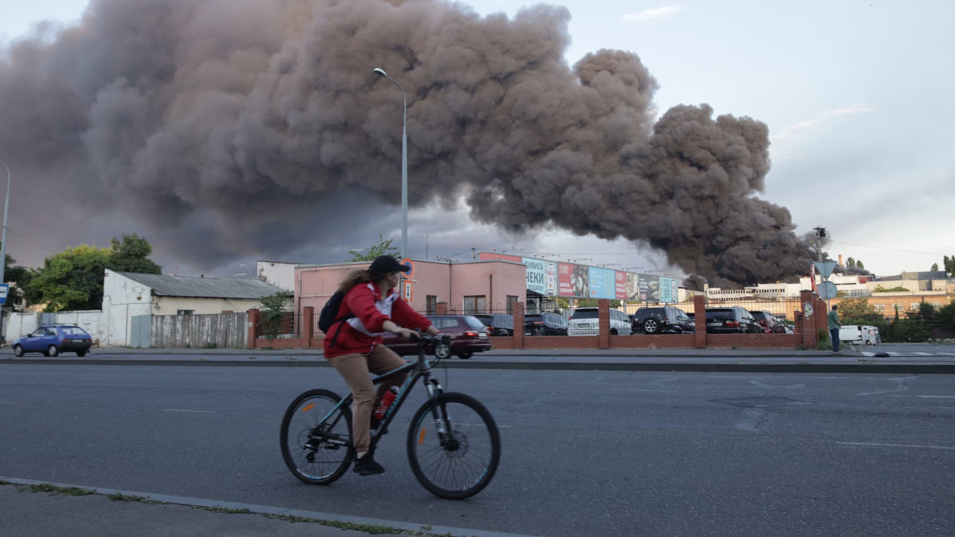 A woman rides a bicycle past a cloud of smoke from a fire in the background, after a missile strike on a warehouse of an industrial and trading company in Odesa on July 16, 2022.