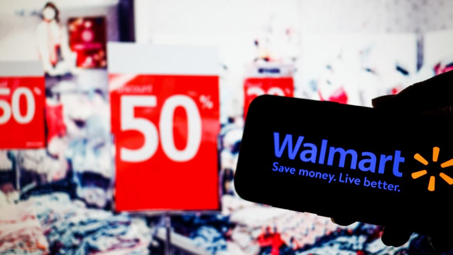 POLAND - 2021/11/24: In this photo illustration, a Walmart logo seen displayed on a smartphone and a department store with a variety of sale banners in the background. (Photo Illustration by Filip Radwanski/SOPA Images/LightRocket via Getty Images)