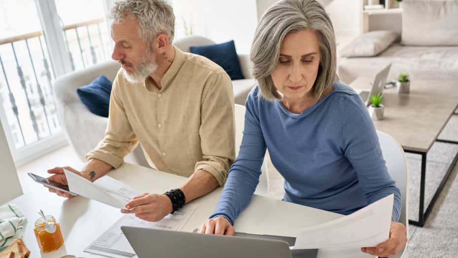 Serious mature senior old couple reading documents bills paying bank loan online together using laptop and phone, calculating pension taxes, planning family retirement money finances working at home.