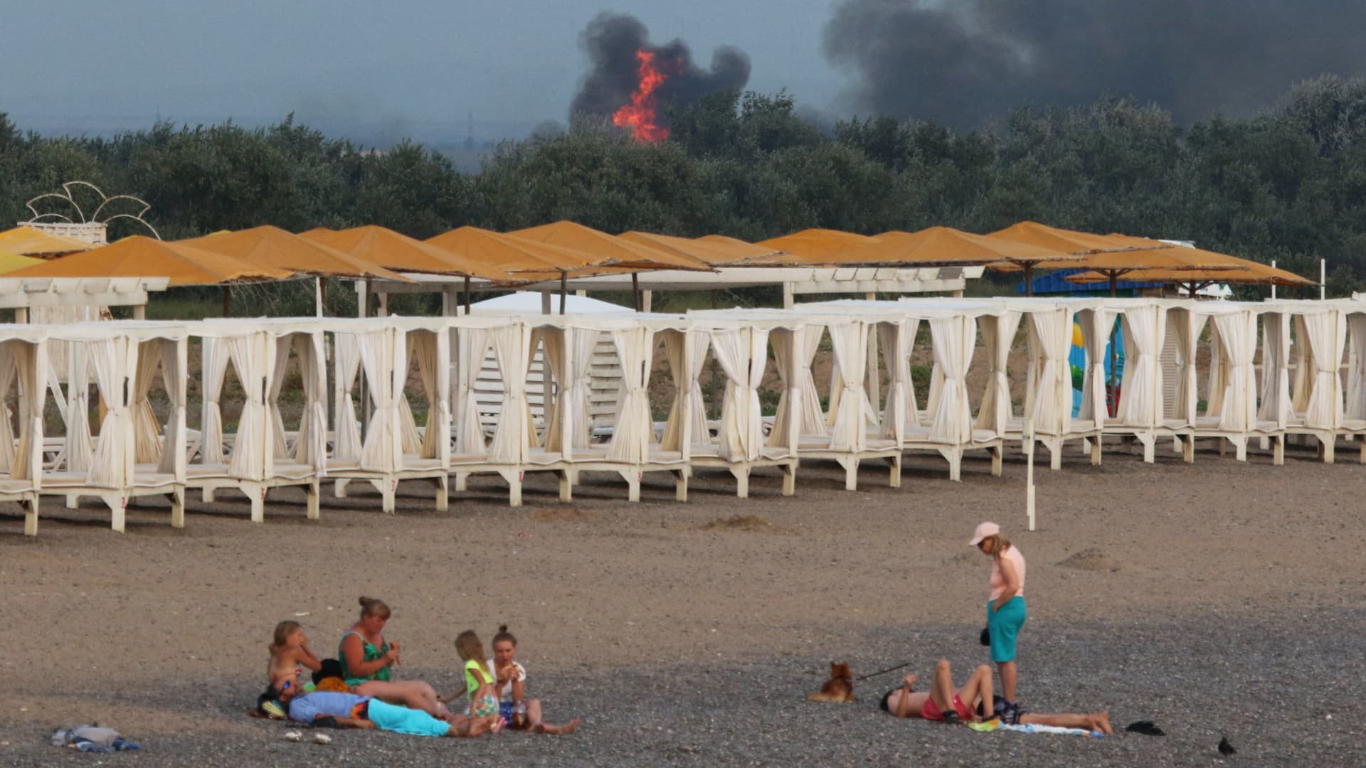 People rest on a beach as smoke and flames rise after explosions at a Russian military airbase, in Novofedorivka, Crimea August 9, 2022. 