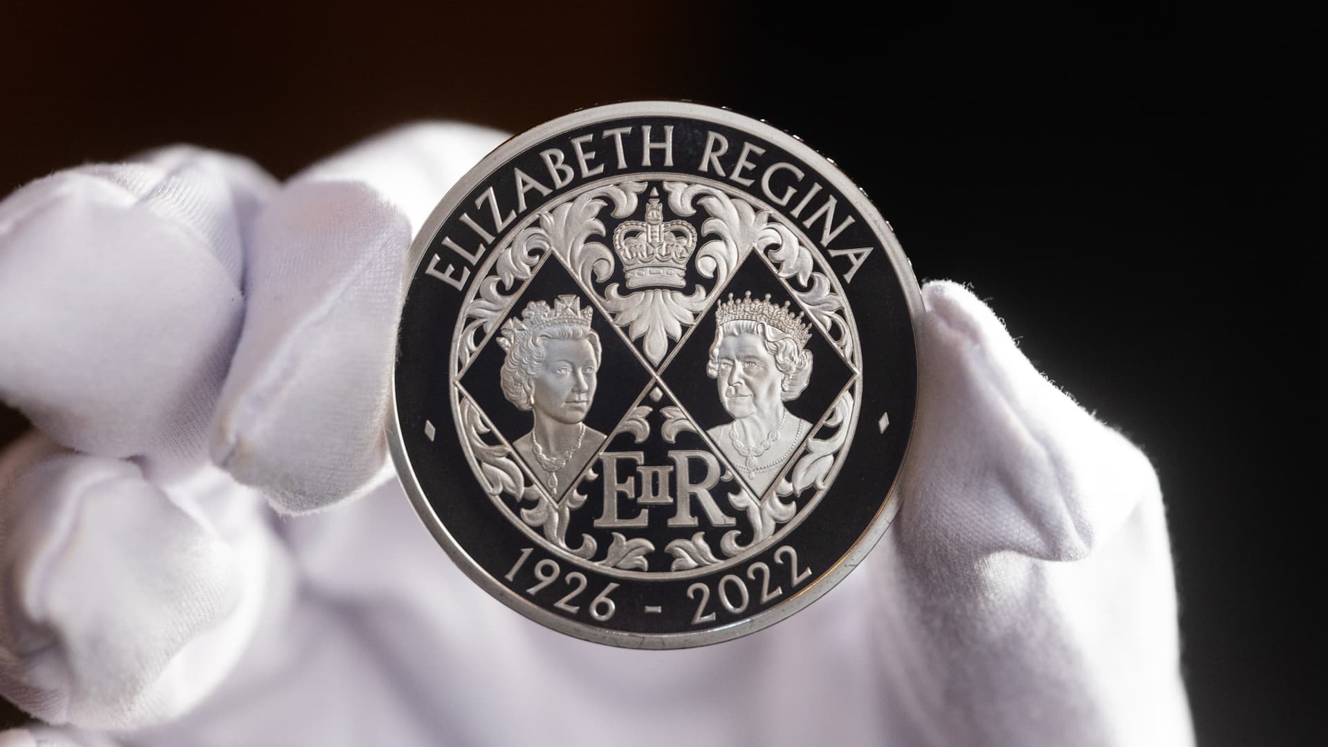 The reverse of a five pound commemorative crown piece coin featuring two portraits of Queen Elizabeth II held by an employee of the Royal Mint.