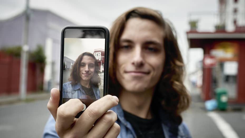 Un joven de pelo largo que usa un teléfono móvil para tomarse un selfie en una calle urbana.