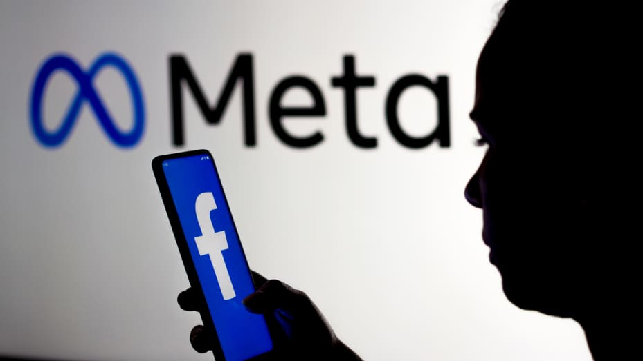 A woman holds a smartphone with the Facebook logo displayed on the screen with the Meta Platforms logo displayed in the background.