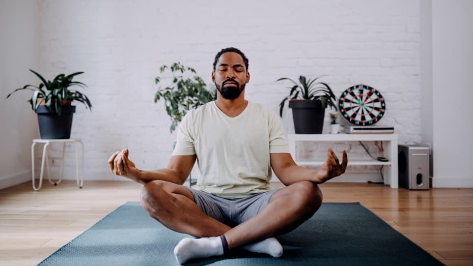 Sporty mid adult black man meditating alone at home, peaceful calm hipster fit guy practicing yoga in lotus pose indoors holding hands in mudra, freedom and calmness concept.