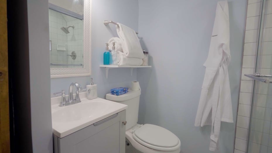 The bathroom features a shelf for extra storage and a glass shower door, which makes the room feel bigger.