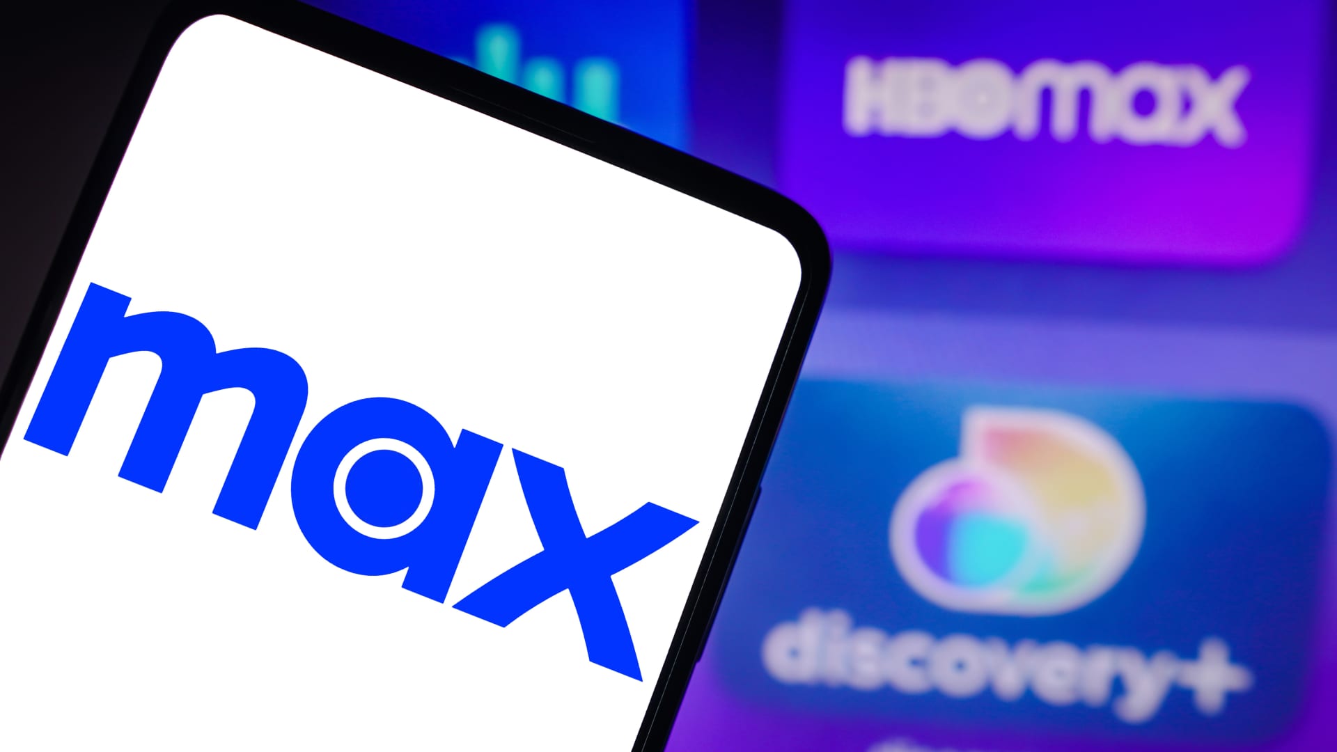 In this photo illustration, the Max logo is seen displayed on a smartphone, the HBO Max and Discovery+ logo in the background. 