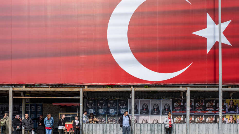 Residentes esperando en una parada de autobús bajo una gran bandera turca en Estambul, Turquía, el domingo 30 de abril de 2023.