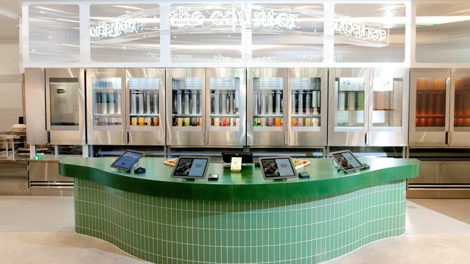 Image shows a counter with four tablets for ordering. Behind the counter are the automated dispensers for Sweetgreen's Infinite Kitchen, holding an array of ingredients.