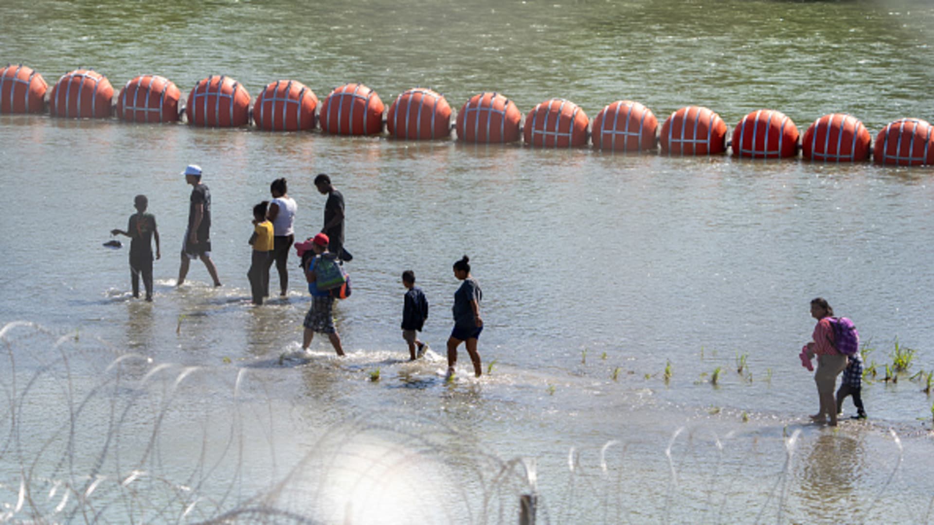 Migrants walk by a string of buoys placed on the water along the Rio Grande border with Mexico in Eagle Pass, Texas, on July 15, 2023, to prevent illegal immigration entry to the US. 