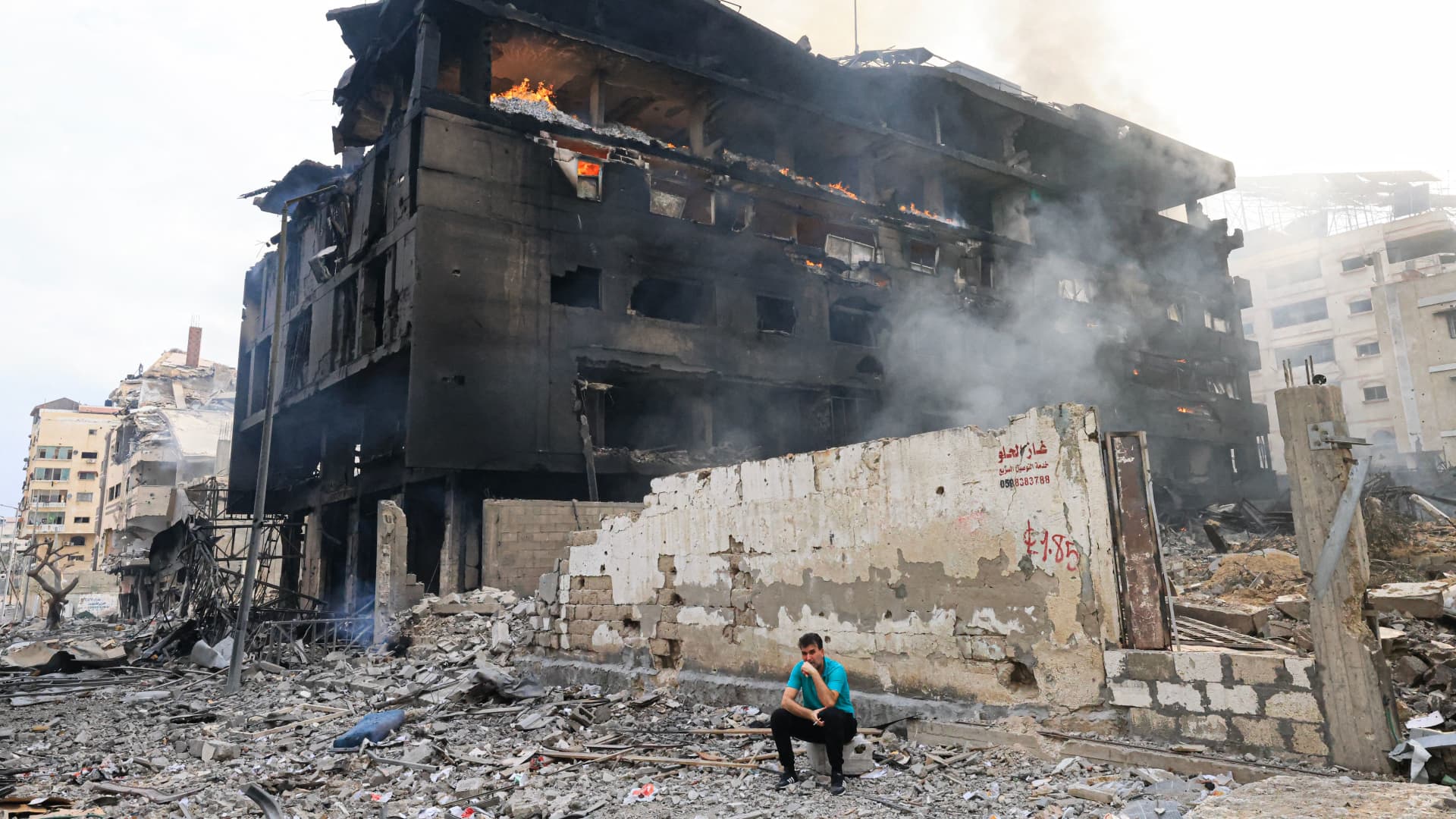 A Palestinian youth sits in front of a charred building as a fire rages through its interior, following Israeli airstrikes in Gaza City's al-Rimal district on October 10, 2023. 