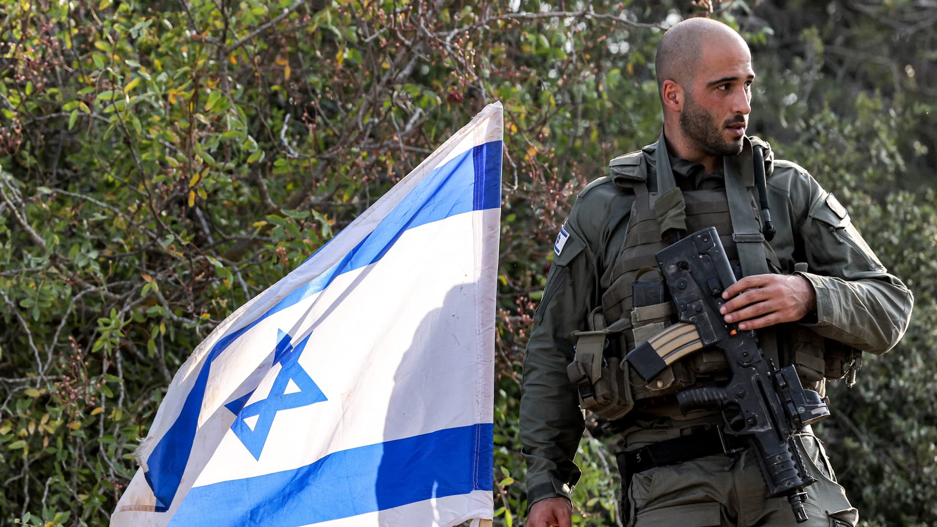 An Israeli army soldier stands with an assault rifle hanging across his chest by an Israeli flag at a position in the upper Galilee region of northern Israel near the border with Lebanon on October 28, 2023 amid increasing cross-border tensions between Hezbollah and Israel as fighting continues in the south with Hamas militants in the Gaza Strip. 