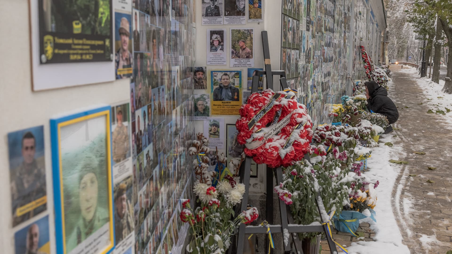 A woman sits in a street blanketed in snow, in front of the memorial wall of fallen defenders of Ukraine in the Russian-Ukrainian war, in downtown Kyiv, on November 22, 2023.