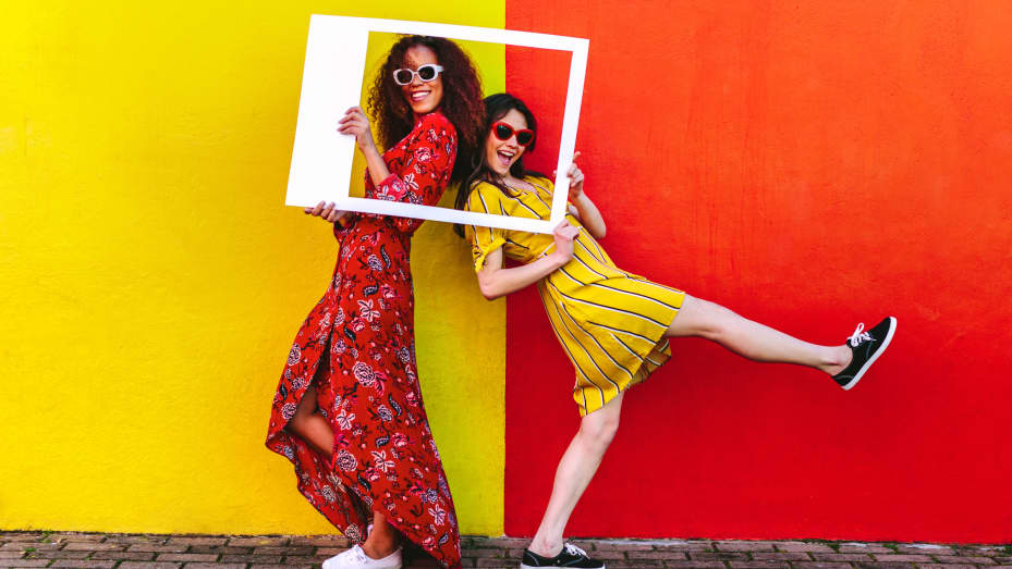 Dos amigas con marco de fotos en blanco de pie contra una pared coloreada al aire libre.  Viajeras femeninas posando ante la cámara con un marco vacío.